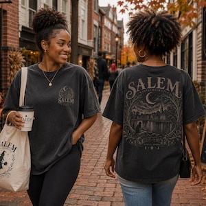 May include: A charcoal gray short-sleeve t-shirt featuring a small Salem graphic on the front left chest and a large, detailed illustration of the Salem waterfront with a crescent moon and cat on the back. A matching cream canvas tote bag is included.