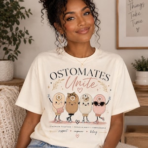 May include: A cream-colored t-shirt featuring the text "OSTOMATES Unite" above a graphic of four cartoon ostomy bags with faces, arms, and legs. Below the characters, the text reads "STRONGER TOGETHER PROUD ALWAYS UNBREAKABLE" and "support empower belong".