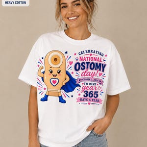 May include: A white short-sleeve t-shirt featuring a cartoon character dressed as a superhero with a blue cape and boots. The graphic includes the text: CELEBRATING NATIONAL OSTOMY day! OCTOBER 3, 2026 I'M IN MY gear 365 DAYS A YEAR.