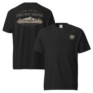 Puede incluir: Una camiseta negra de manga corta con un pequeño logotipo hexagonal C&F en el pecho. La parte posterior muestra un gráfico de una cordillera con pinos y el texto MADE FOR THE LONG WAY AROUND CANYON & FORGE EST. 2025 en beige y blanco.