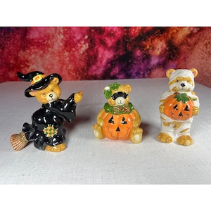 May include: Three ceramic bear figurines dressed for Halloween. The first is a bear in a black witch costume with a broom. The second is a bear wearing a black mask inside a carved orange pumpkin. The third is a bear wrapped in white mummy bandages holding a pumpkin.