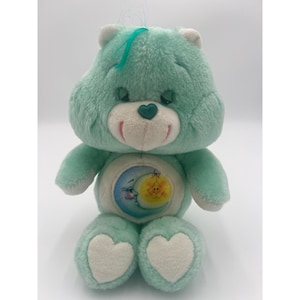 May include: A mint green plush bear toy with a white muzzle and a dark green heart-shaped nose. The bear features a circular patch on its belly depicting a blue crescent moon and a yellow star. It has white heart-shaped patches on its feet.
