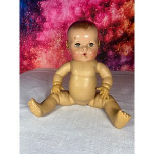 May include: A vintage baby doll with a molded plastic body, light skin tone, and painted facial features. The doll has brown hair, green eyes, and an open mouth. It is posed in a seated position with arms and legs extended outward, showcasing a smooth, jointed construction typical of mid-century playthings.