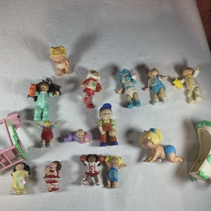 May include: A collection of small plastic Cabbage Patch Kids figurines including various characters in different outfits, a pink plastic swing set, and a white cradle with green accents. The figures feature diverse hair colours, clothing styles, and poses.