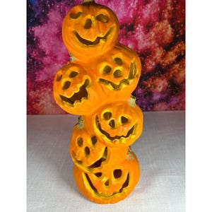 May include: A vertical stack of five orange ceramic jack-o'-lanterns with carved facial features including triangular eyes and jagged mouths. Each pumpkin is connected to the next, creating a single decorative pillar with a bright finish.