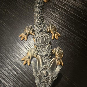 May include: A 3D printed articulated dragon figurine featuring a dark grey body with metallic gold accents on the claws, head, and tail tip. The creature has a segmented, scale-like design with a detailed head, four legs, and a long, flexible tail.