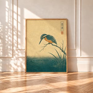 May include: A framed wall art print featuring a kingfisher bird perched on a branch above dark teal water with ripples. The background is a textured light yellow. The print includes vertical Japanese text and a red square seal stamp.