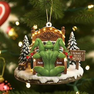 Peut inclure: Une décoration de Noël représentant une créature verte en talons hauts rouges agenouillée sur un cadre de lit. Le lit porte les noms David et Susan. Un panneau en bois à côté du lit indique I licked it So it's MINE. La scène inclut deux sapins.