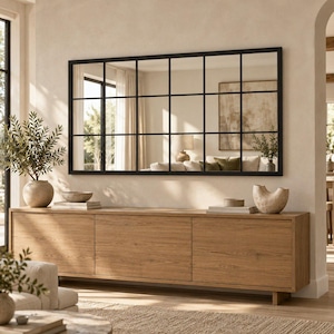 May include: A large rectangular wall mirror featuring a black grid frame with fifteen individual panes. The mirror is mounted horizontally above a long, light-toned wooden sideboard cabinet with three cabinet doors and a smooth, flat surface.