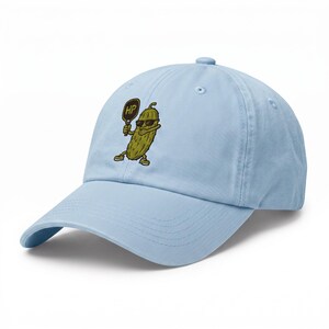 May include: A light blue baseball cap featuring an embroidered graphic of a green pickle wearing black sunglasses and holding a brown pickleball paddle with the letters HP printed on it. The cap has a curved brim and a button on the top.
