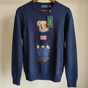 May include: A navy blue long-sleeve crewneck sweater featuring a knit graphic of a teddy bear wearing a sweater with an American flag design and blue jeans. The sweater has a Polo Ralph Lauren tag attached to the collar and red RL embroidery.