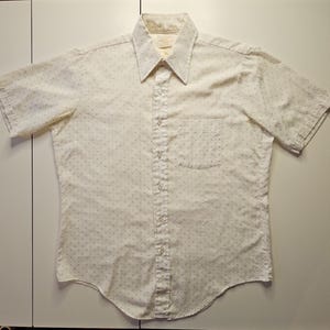 May include: A short-sleeved button-down shirt in an off-white color featuring a subtle, repeating pattern of small, dark-colored dots. The shirt includes a pointed collar, a front button placket, and a single chest pocket on the left side.