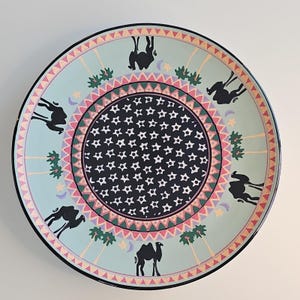 May include: A round ceramic plate featuring a central black circle filled with white star patterns. The border displays a light blue background with repeating black camel silhouettes, green palm trees, yellow stars, and crescent moons, framed by a pink and green triangular pattern.