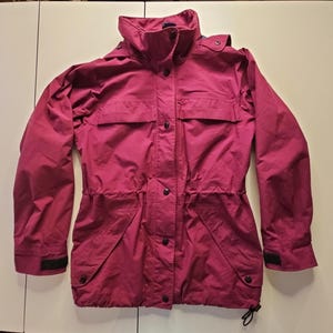May include: A magenta long-sleeve jacket featuring a high collar, a front zipper closure concealed by a snap-button placket, two chest pockets, two lower pockets with snap closures, a drawstring waist, and adjustable hook-and-loop wrist cuffs.