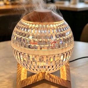 May include: A spherical ultrasonic humidifier with a faceted, clear glass-like body and a wood-grain top. It emits a steady stream of mist from the centre. The device features the text Digital Bazaar 24 and a red circular logo, resting on a wooden stand.