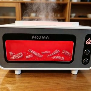 May include: A white and grey rectangular aroma diffuser with a front window displaying a red glowing fire effect with simulated embers. The word AROMA is printed above the window, and a control panel with three icons is on the right side.