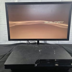 May include: A black PlayStation 3 game console sits in front of an ASUS computer monitor. The monitor displays a brown screen with the text "EVILNAT" in white lettering. The console has a rectangular shape with a disc slot on the front panel.
