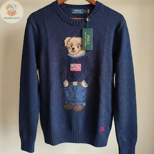 May include: A navy blue long-sleeve crewneck sweater featuring a knit graphic of a teddy bear wearing a sweater with an American flag design and blue jeans. The sweater has a Polo Ralph Lauren brand tag attached and the letters RL in red.