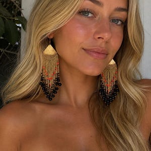Puede incluir: Un par de pendientes largos y colgantes con una pieza superior triangular de tono dorado. Debajo del triángulo, múltiples hilos de cuentas doradas están acentuados con pequeñas cuentas de color naranja brillante y terminan con cuentas negras redondas.