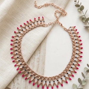 Puede incluir: Un collar de cuentas con un patrón geométrico de celosía hecho de cuentas de semillas en color tostado, negro y rosa brillante. El collar incluye una cadena de metal en tono oro rosa con cierre de mosquetón y un diseño estructurado.