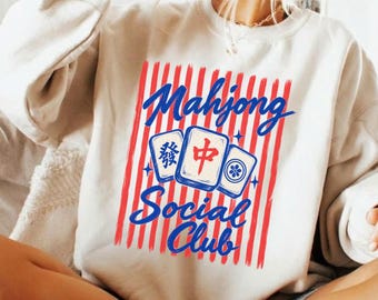 May include: A cream-coloured crewneck sweatshirt featuring a graphic print of red vertical stripes. Over the stripes, three mahjong tiles are displayed with blue text that reads Mahjong Social Club in a cursive script font.
