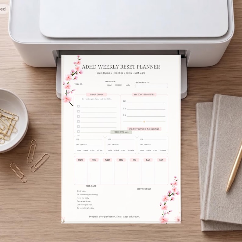 May include: A white paper ADHD weekly reset planner featuring pink cherry blossom illustrations on the top left and bottom right corners. The layout includes sections for brain dump, priorities, tasks, self-care, and a daily schedule for the week.