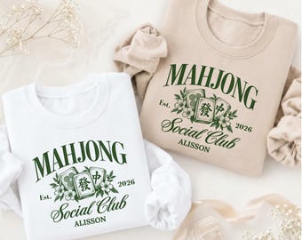 May include: A white and a beige crewneck sweatshirt, each featuring a dark green graphic with the text "MAHJONG SOCIAL CLUB", "Est. 2026", and "ALISSON", accompanied by illustrations of mahjong tiles and small floral accents.