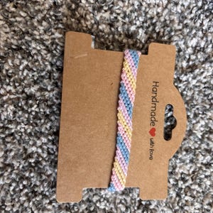May include: A woven friendship bracelet featuring a diagonal striped pattern in pastel pink, light blue, and pale yellow threads. The bracelet is attached to a rectangular brown cardboard display card with the text Handmade with love.