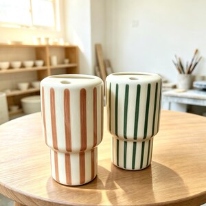 May include: Two cylindrical ceramic vases with a stepped design. One vase features vertical terracotta-colored stripes on a cream background, while the other features vertical dark green stripes on a cream background. Both have a small circular opening.