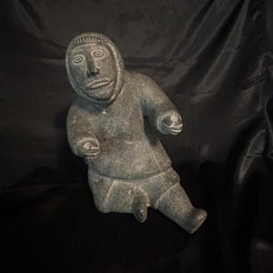 Peut inclure: Une sculpture en pierre grise représentant un personnage assis portant une parka à capuche. La figurine présente des traits du visage sculptés, notamment les yeux, le nez et la bouche, avec les bras tendus vers l'avant et les jambes assises.