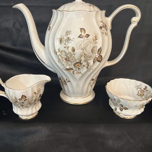 May include: A white porcelain teapot with a matching creamer and sugar bowl, featuring a fluted body design, gold trim on the edges, and a floral pattern in shades of brown and tan on the exterior of each piece, including a lid on the teapot.
