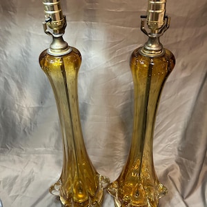 Op de afbeelding: Een paar amberkleurige glazen tafellampen met een slanke, langwerpige hals en een uitlopende, gegolfde voet. Elke lamp is voorzien van een goudkleurige metalen fitting aan de bovenzijde voor een lichtbron, met een lichaam van helder glas.