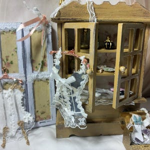 May include: A miniature wooden cabinet with open doors, a folding screen with floral panels, and two small garments on copper-colored hangers. The cabinet features shelves holding tiny decorative items, while a small box contains lace accessories.