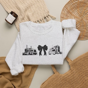 May include: A light gray crewneck sweatshirt featuring a black graphic print of two semi-trucks positioned on either side of a large black bow. The long-sleeved garment has a ribbed collar, cuffs, and hem, showcasing a clean and simple design.