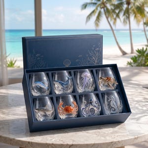 Op de afbeelding: Een set van acht heldere, steelvrije wijnglazen in een donkerblauwe presentatiedoos. Elk glas is voorzien van een uniek driedimensionaal zeedierfiguur, waaronder een dolfijn, walvis, kwal, zeeschildpad, haai, krab, octopus en zeepaardje.