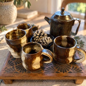 Può includere: Un set di tre tazze in ceramica e una teiera abbinata con smalto maculato marrone scuro e crema. Gli oggetti sono disposti su un vassoio rettangolare in legno con un motivo a ventaglio intagliato, con una ciotola di pigne al centro.