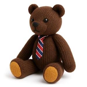 Op de afbeelding: Een bruine gebreide teddybeer met een geribbelde textuur, zwarte plastic ogen en een donkere neus. De beer draagt een das met diagonale strepen in rood, wit en marineblauw. De onderkant van de voeten is uitgevoerd in contrasterend mosterdgeel.