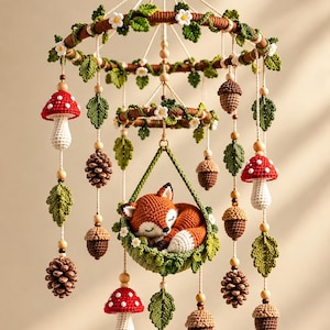 May include: A hanging mobile featuring a crocheted sleeping fox in a green basket, surrounded by crocheted red and white mushrooms, brown acorns, pinecones, and green leaves with small white flowers, all suspended from two circular wooden rings.