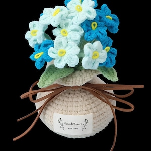 May include: A crochet flower arrangement featuring light blue and dark blue blossoms with yellow centers and green leaves. The flowers are held in a round, cream-colored crocheted pot tied with a brown leather cord and a label reading Handmade WITH LOVE.
