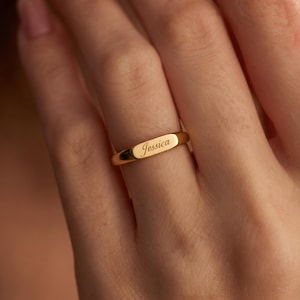 Puede incluir: Un anillo de tono dorado con una placa central plana y rectangular grabada con el nombre Jessica en letra cursiva. El anillo tiene una superficie pulida y reflectante, con una banda delgada y uniforme que rodea el dedo.