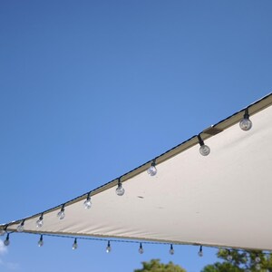 Könnte beinhalten: Ein dreieckiges, cremefarbenes Sonnensegel, das vor einem klaren blauen Himmel gespannt ist. Eine Lichterkette mit kleinen, runden, klaren Glühbirnen ist entlang der Stoffkante befestigt und bildet einen dekorativen Rand vor dem hellen Hintergrund.