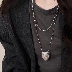 May include: A silver-toned double-layered necklace featuring a heart-shaped locket pendant with intricate embossed floral patterns. The necklace is worn over a dark gray t-shirt and a black cardigan, showcasing a delicate chain design.