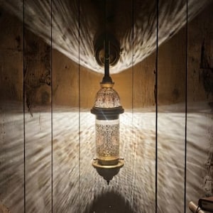 May include: A wall-mounted lantern with a cylindrical body featuring intricate metal cutouts. The fixture emits a warm, glowing light that projects a distinct, rippled pattern of shadows and illumination onto the surrounding vertical surface.