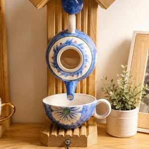 May include: A blue and white ceramic teapot and teacup set mounted on a light wood structure. The teapot is suspended above the teacup, which features a floral pattern. A black metal chain hangs from the base of the wooden mount.