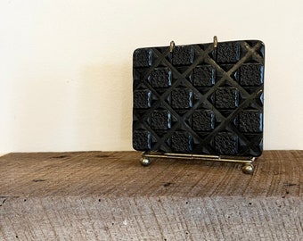 May include: A black, square decorative object with a geometric pattern, displayed on a small gold-colored metal stand. The object has a repeating diamond and floral design. The stand has a small ball on each foot.