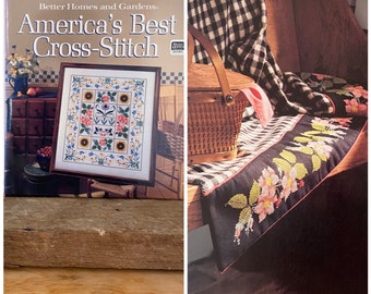 May include: A book titled "Better Homes and Gardens America's Best Cross-Stitch" is displayed next to a framed cross-stitch design. A picnic basket and a black table runner with floral embroidery are also visible.