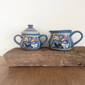 May include: A blue ceramic sugar bowl with a lid and a matching creamer. Both pieces feature a floral design in yellow, red, and black. The sugar bowl has two handles, and the creamer has one handle. The set is displayed on a rustic wooden surface.