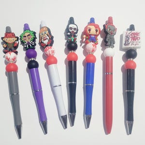 May include: A set of seven pens featuring horror-themed character toppers and decorative beads. The pens have barrels in grey, purple, white, black, blue, and red. One pen features a square white topper with the text One Two Freddy's coming for you.