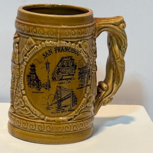 Könnte beinhalten: Ein beigefarbener Bierkrug aus Keramik mit einem reliefartigen Henkel und einem zentralen kreisförmigen Motiv mit der Aufschrift San Francisco. Das Design zeigt schwarze Strichzeichnungen einer Seilbahn, Chinatown, Fisherman's Wharf und der Golden Gate Bridge.