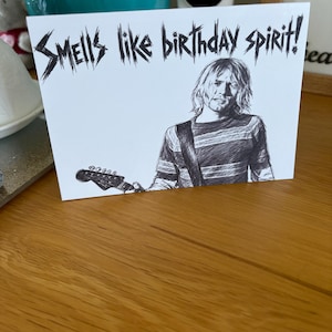 May include: A white rectangular greeting card featuring a black ink illustration of a musician with long hair wearing a striped shirt and holding a guitar. The text "SMELLS like birthday spirit!" is printed in a jagged, stylised black font.